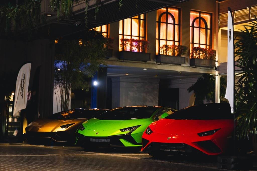 Shiny luxury sports cars line a vibrant Bangkok street at night as shoppers look on.