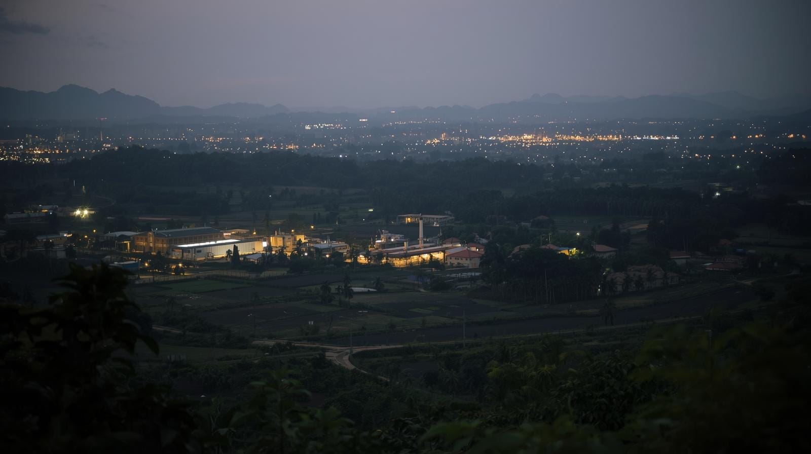 Dusk view over a Thai provincial area showing a mix of small factories, fields and a distant refugee camp, with lights turning on across the landscape.